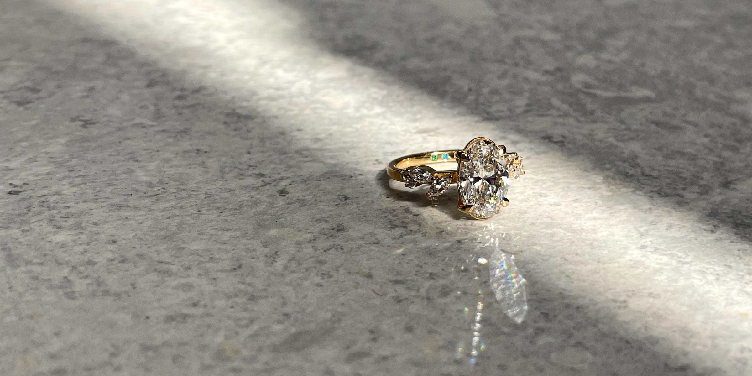 Custom, ethical and sustainable engagement ring with oval labgrown diamond and marquise side stones in recycled yellow gold photographed in natural light on marble. Nature inspired bespoke design with solitair ring for the perfect proposal ring.