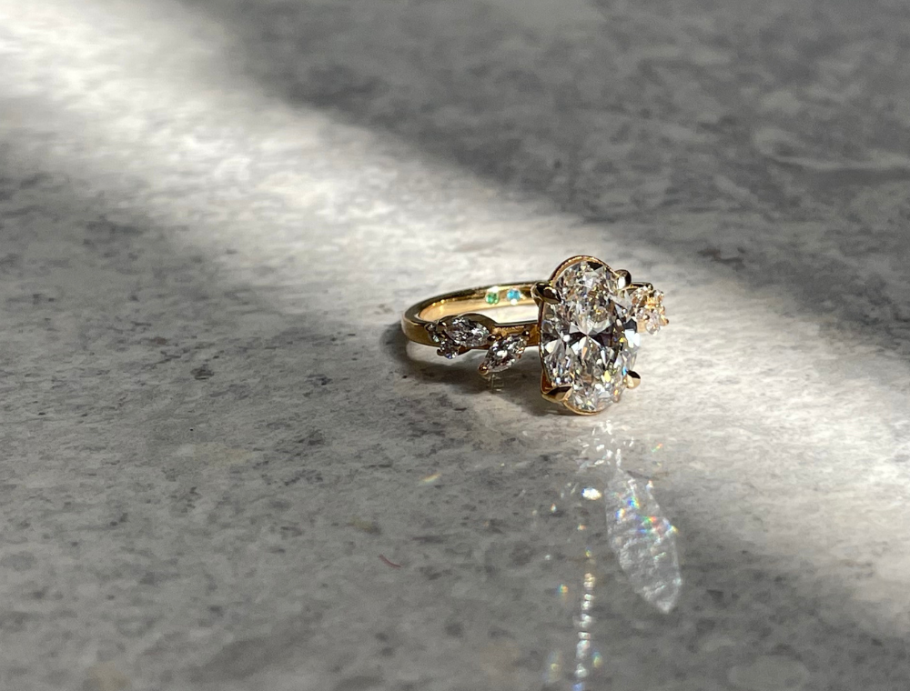 Custom, ethical and sustainable engagement ring with oval labgrown diamond and marquise side stones in recycled yellow gold photographed in natural light on marble. Nature inspired bespoke design with solitair ring for the perfect proposal ring.