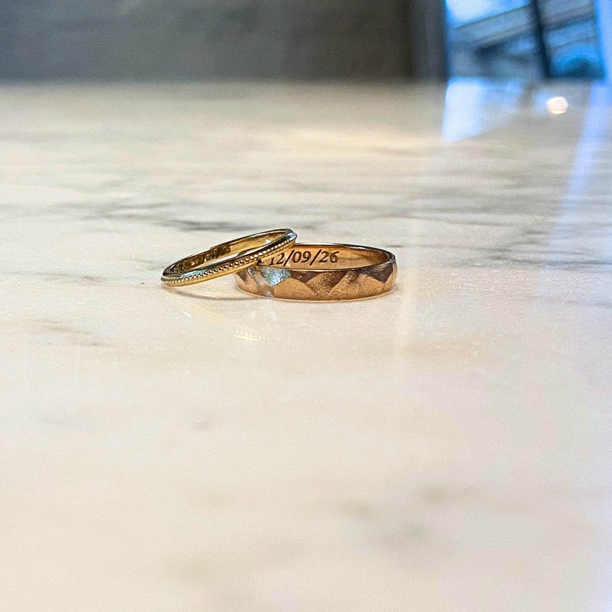Laurel Ring, a beaded gold wedding band, beside an engraved hammered band on marble, recycled 18K gold