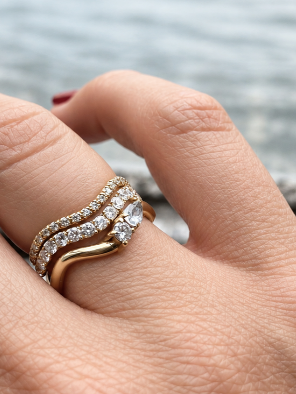 A close-up of a hand wearing a stack of three gold wave rings set with labgrown diamonds, including two pavé diamond bands and a gold band with a pear-cut diamond and a brilliant-cut diamond.