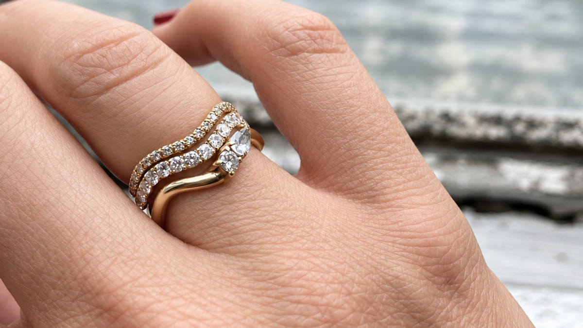 A close-up of a hand wearing a stack of three gold wave rings set with labgrown diamonds, including two pavé diamond bands and a gold band with a pear-cut diamond and a brilliant-cut diamond.