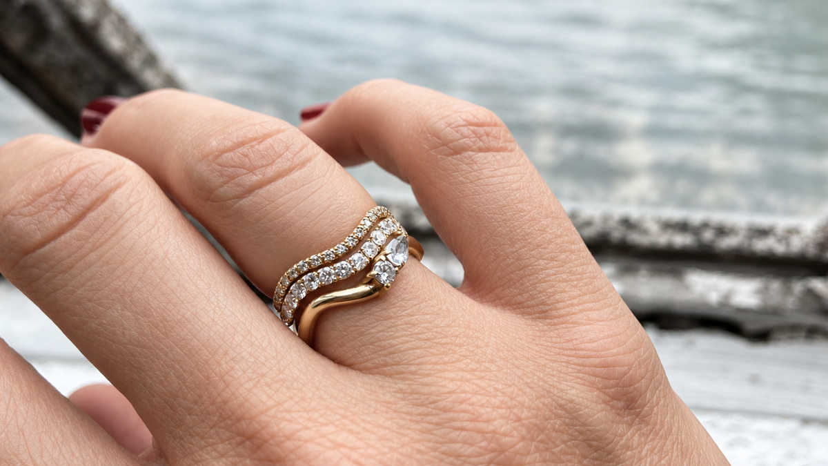 A close-up of a hand wearing a stack of three gold wave rings set with labgrown diamonds, including two pavé diamond bands and a gold band with a pear-cut diamond and a brilliant-cut diamond.