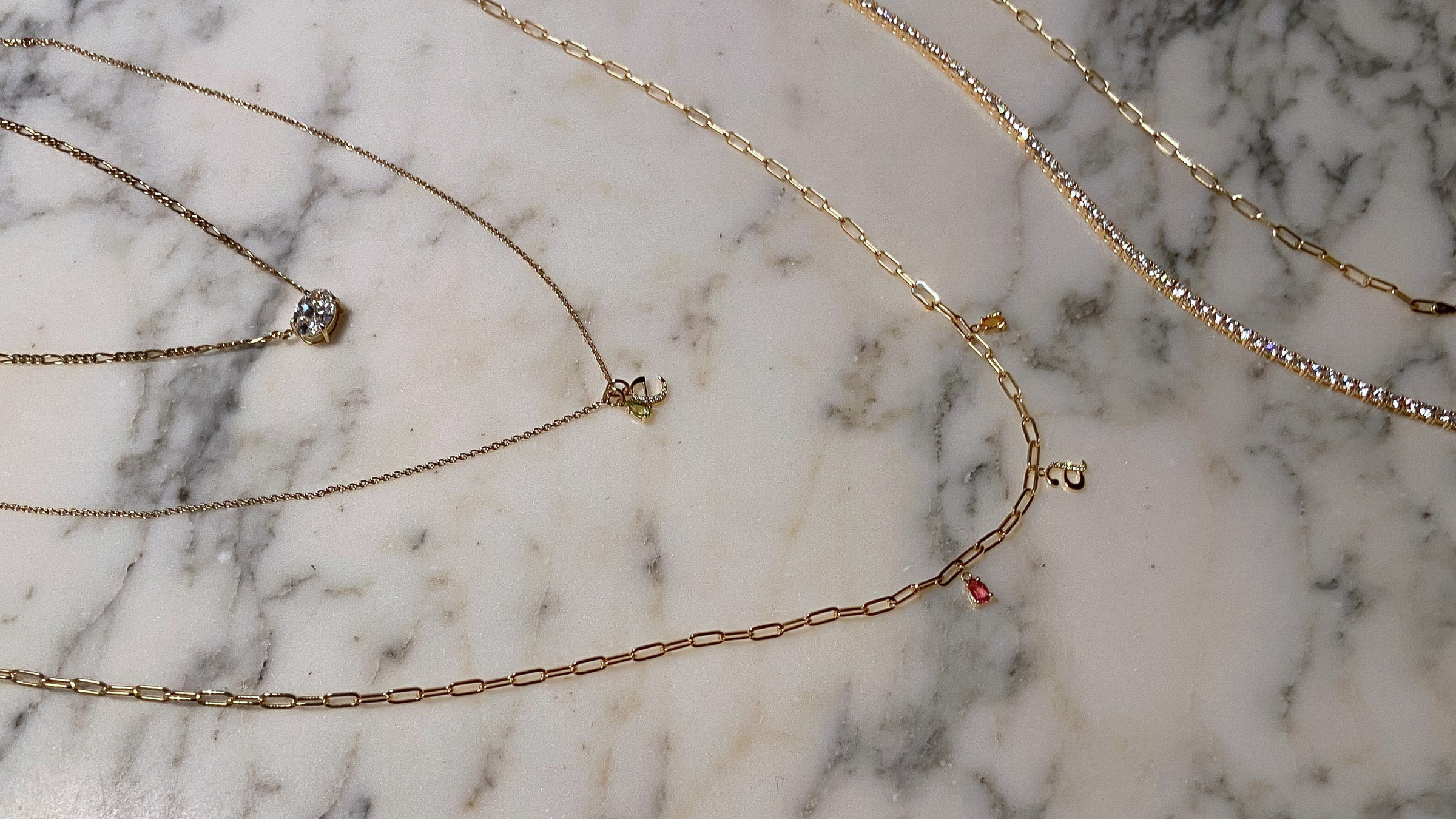 Dainty gold layered necklaces with labgrown diamonds, chain links, initials pendants, birthstone charms, and labgrown diamond tennis bracelets displayed on marble background.