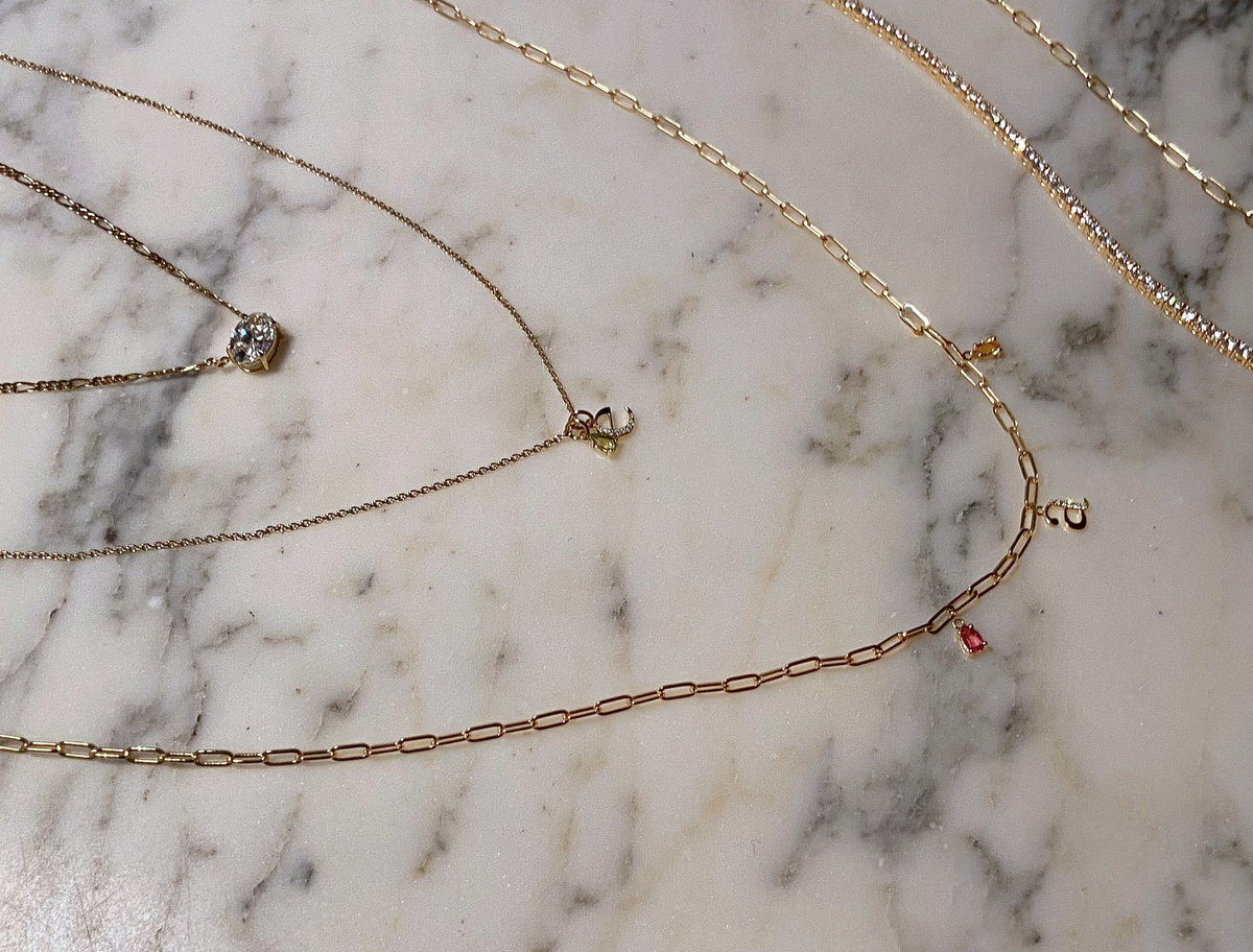 Dainty gold layered necklaces with labgrown diamonds, chain links, initials pendants, birthstone charms, and labgrown diamond tennis bracelets displayed on marble background.